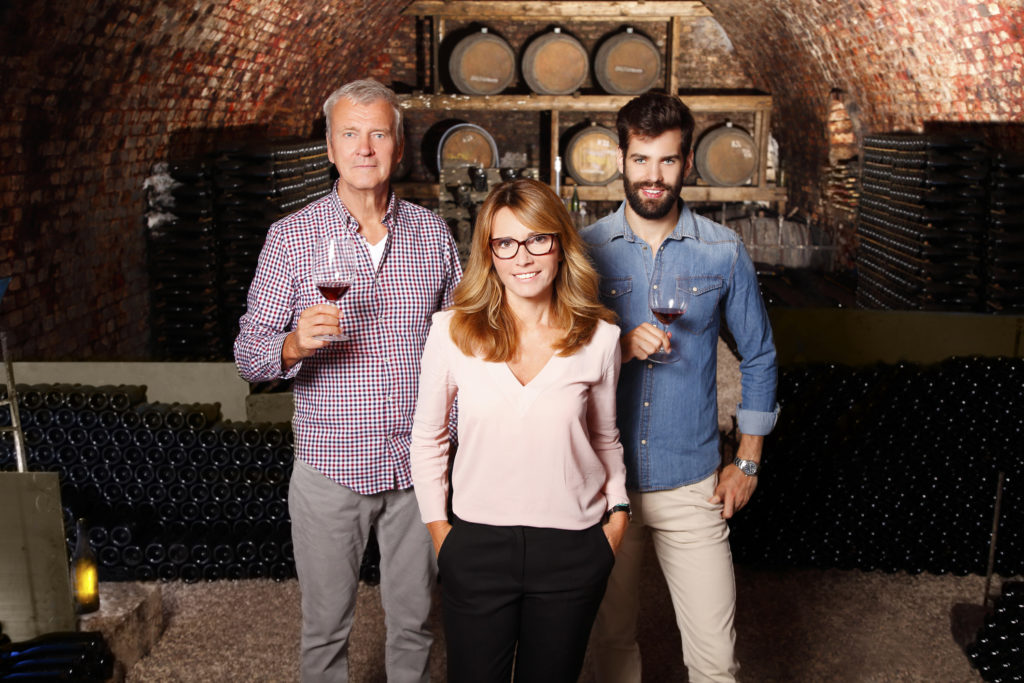 Portrait of multigenerational winery owner family standing at wine cellar. Senior winemaker and young sommelier standing at background and holding in hands a glass of red wine while middle age businesswoman looking at camera and smiling. Small business.