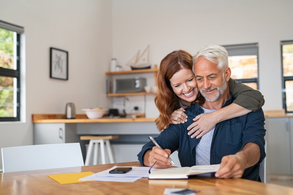 Loving mature wife embracing husband from behind while writing in book. Happy middle aged couple making to do list of purchases and discussing future plans. Cheerful senior man working at home on wooden table with beautiful woman hugging him from behind, copy space.